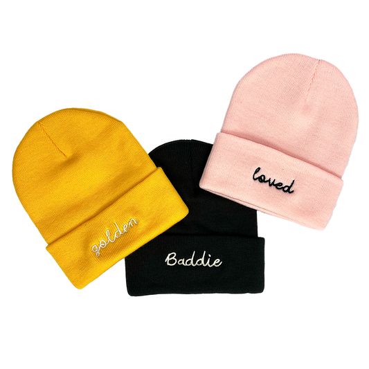 Three beanies in yellow, black, and pink with embroidered text on a white background